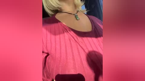 Media: Video of a person wearing a loose, pink, ribbed blouse with a small, green pendant necklace. The background is blurred, with a pink overlay.