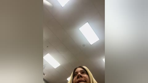 Media: A video of a low-angle view from a tall person looking up, revealing a blonde woman's face, ceiling lights, and a white wall.