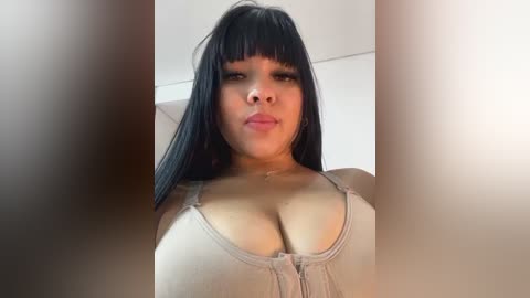 Media: Video of a young Latina woman with long black hair and bangs, wearing a beige tank top that highlights her ample cleavage. The background is a plain white wall.