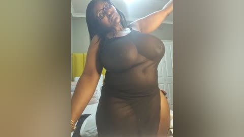 Media: Video of a curvy Black woman with long hair, wearing a sheer black dress, standing in a bedroom with white bed and yellow wall.