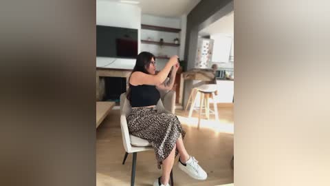 Media: A video of a woman in a modern, minimalist living room. She sits on a gray chair, wearing a black crop top and leopard print skirt, adjusting her glasses. The room features light wooden floors, a white fireplace, and a kitchen with a bar.