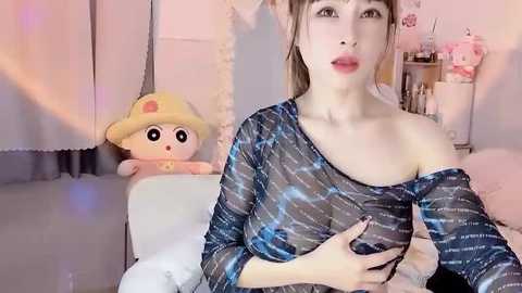 Media: Video of a slender, fair-skinned woman with brown hair, wearing a sheer, blue-striped off-shoulder top, sitting on a white couch. Behind her is a plush toy and a soft, pink background.