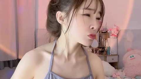 Video of a young Asian woman with pale skin, dark hair in a ponytail, wearing a light blue halter top, blowing a kiss, in a softly lit, pastel-colored bedroom with stuffed toys.