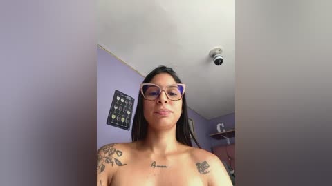 Media: A video of a topless woman with long dark hair, wearing glasses and multiple tattoos, including \"A\" on her chest, standing in a room with lavender walls, a clock, and a dark framed picture.