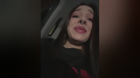 Media: Video of a young woman with long brown hair, light skin, and a septum piercing, wearing a black hoodie. She appears slightly out of focus, with a dark background, creating a moody atmosphere.