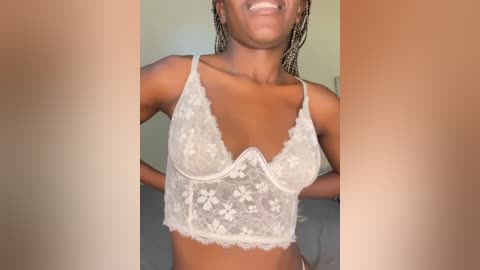 Media: Video of a smiling Black woman with dark skin and braided hair, wearing a white lace bralette, indoors with blurred background.