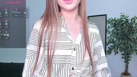 Media: Video of a light-skinned woman with long, straight, reddish-brown hair, wearing a white and black striped button-down shirt. Background includes a green plant and a black-framed wall calendar.