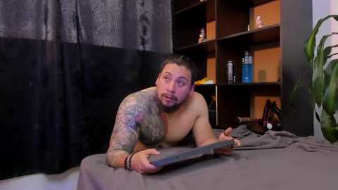 Media: Video of a shirtless, muscular, tattooed man with a scruffy beard, lying on a bed, holding a tablet. Background features a dark curtain, wooden shelves, and a potted plant.