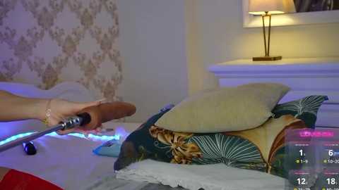 Media: Video of a woman holding a wooden spoon over a bed with a green leaf-patterned blanket and pillows. The room has a floral wallpaper and a beige lamp on a white dresser.