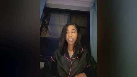 Media: Video of a young African American woman with medium skin tone and long, straight black hair, wearing a black satin robe with pink trim, standing in a dimly lit hallway.