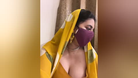 Media: Video of a South Asian woman with medium brown skin, wearing a mustard yellow saree with green and white accents, a purple face mask, and gold earrings. She is indoors with beige curtains in the background.