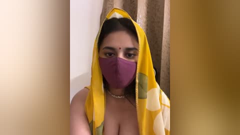 Media: Video of a South Asian woman with medium skin tone, wearing a yellow sari with white polka dots, purple face mask, and a bindi, set against beige curtains.