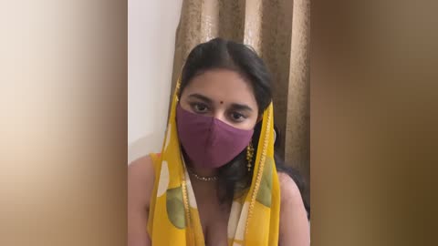 Media: Video of a South Asian woman with medium skin tone, wearing a yellow sari with green patterns, a purple face mask, and a red bindi, standing in front of beige curtains.