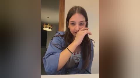 Media: Video of a young woman with long brown hair, wearing a blue hoodie, standing in front of a mirror, covering her mouth with her hand, while a crown is visible on the wall behind her.