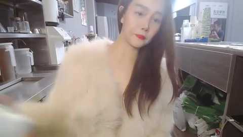 Media: Video of an Asian woman with long brown hair, wearing a white fur coat, standing in a cluttered kitchen with stainless steel appliances, green groceries, and a partially open refrigerator.