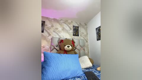 Media: A video of a cozy bedroom with a large teddy bear in the center, surrounded by a bed with blue and white bedding, a white wall with abstract art, and a pink light on the ceiling.