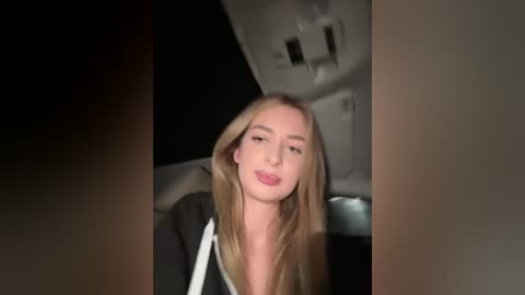 Media: A video of a blonde woman with long hair, fair skin, and a slim physique, wearing a black jacket, sitting in a car with a dark interior.