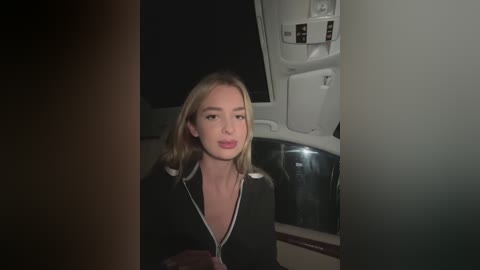 Media: Video of a blonde woman with straight hair, wearing a black jacket, standing in a dimly lit room with a white sink and a dark background.