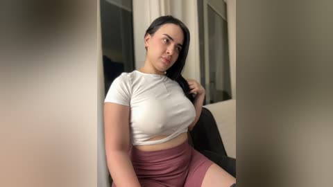 Media: Video of a curvy, light-skinned woman with long black hair, wearing a tight white crop top and high-waisted maroon shorts, seated on a black chair, indoors.