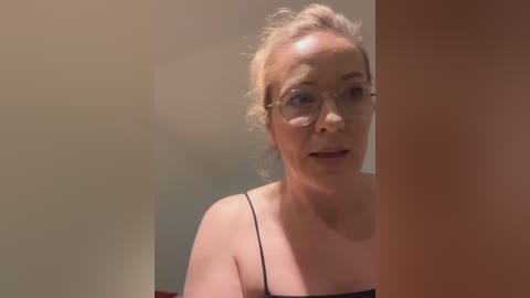 Media: A video of a middle-aged woman with fair skin, blonde hair tied back, wearing glasses and a black spaghetti-strap top. She's speaking with an expression of concern. Background is blurred.