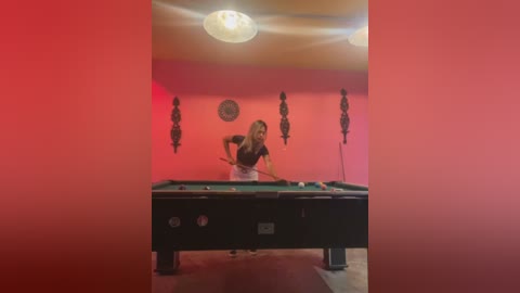 Media: Video of a woman in a red-painted room, wearing a black shirt, standing behind a pool table, with a pool cue in her hand, surrounded by decorative black masks on the wall.