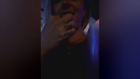 Media: Video of a person with medium skin tone, wearing a black shirt, with a close-up of their hand covering their mouth, hinting at a whisper or a secret. Background is dimly lit, with a blue and purple hue, suggesting an intimate or clandestine setting.