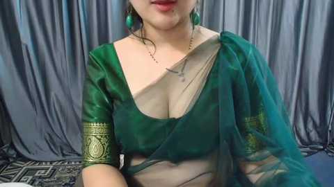 Media: Video of a light-skinned woman with a medium build, wearing a green sari with gold embroidery, green earrings, and a necklace, against a grey curtain backdrop.