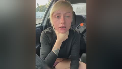 Media: Video of a fair-skinned woman with blonde hair, wearing a black long-sleeve top, resting her chin on her hand, looking contemplative, inside a car.