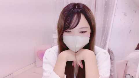 Media: Video of a young East Asian woman with long, straight brown hair, wearing a white mask and white lace top, sitting on a bed with pale pink sheets, looking down.