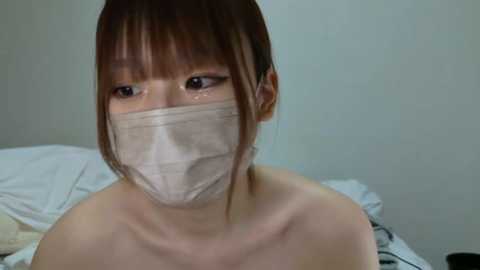 Media: Video of a young East Asian woman with short brown hair and bangs, wearing a beige surgical mask, sitting on a bed with white sheets in a minimalistic, possibly bedroom setting.