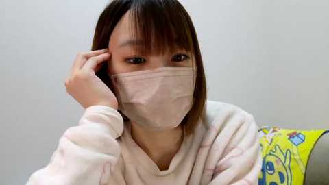 Media: Video of an East Asian woman with straight, shoulder-length brown hair, wearing a white, long-sleeved pajama top, a pink face mask, and a yellow SpongeBob SquarePants pillow. She is touching her hair with her left hand, against a plain white background.