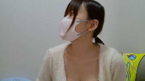 Media: A video of a young Asian woman with light skin, wearing a white mask and light beige cardigan, sitting indoors with a yellow towel visible behind her.