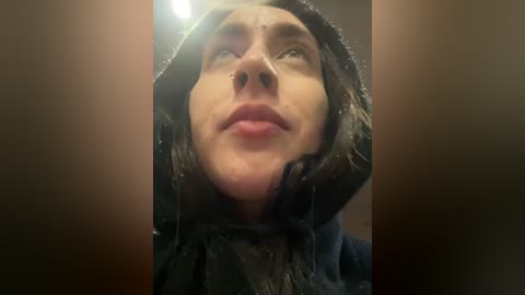 Media: Video of a young woman with wet hair, wearing a black hooded jacket, gazing upward with a contemplative expression. Background is out of focus.