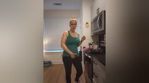 Media: A video of a blonde woman in a teal tank top and black pants standing in a modern kitchen, holding a phone, with white blinds and wooden floors in the background.