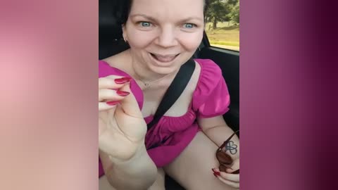 Media: Video of a middle-aged, fair-skinned woman with short black hair, wearing a pink top and black seatbelt, sitting in a car. She has a tattoo on her right hand, red nails, and a relaxed expression.