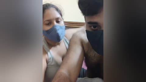 Media: Video of a woman with brown hair, wearing a blue face mask and gray tank top, and a man with dark hair and a black mask, both in a dimly lit room.