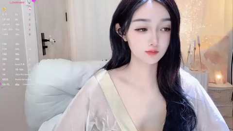 Media: Video of a young Asian woman with pale skin, long black hair, and delicate features, wearing a sheer white top over a black bralette, sitting indoors. Background features a white door and a lit candle.