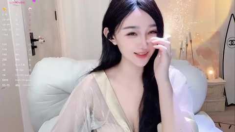 Media: Video of a fair-skinned, slender young woman with long black hair, smiling, wearing a sheer, off-shoulder top, sitting on a white couch, with a soft, beige wall and a lit candle in the background.