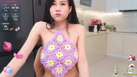 Media: Video of an Asian woman with long black hair, wearing a pink crochet bikini top with white and yellow flowers, in a modern kitchen with white cabinets, black appliances, and a refrigerator.