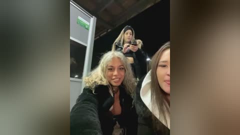 Media: Video of three women in a dimly-lit outdoor setting; one blonde with curly hair, another with straight hair, and a third partially visible, all smiling.