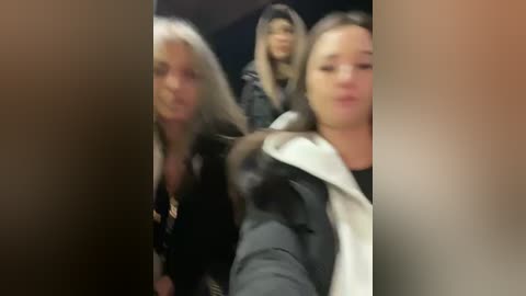 Media: A blurry video shows three women with long hair, wearing black jackets and white scarves, standing close together in an indoor setting.