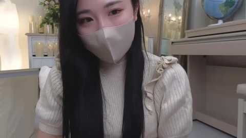 Media: Video of an East Asian woman with long black hair, wearing a beige mask and a beige ribbed sweater with a black scarf, standing indoors with a white door and a potted plant in the background.