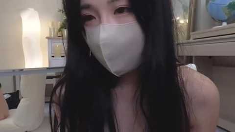 Media: A video of an Asian woman with long black hair and a white surgical mask, wearing a sleeveless top, indoors near a white table with potted plants and a glass aquarium.