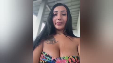 Media: Video of a curvy, light-skinned woman with long black hair, wearing a colorful, strapless top, standing indoors with a metallic ceiling and blurred background.