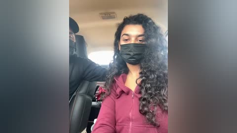 Media: A video of a woman with curly black hair, wearing a black face mask and a maroon hoodie, seated in a car, with a uniformed driver visible in the background.