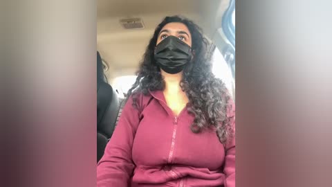 Media: Video of a South Asian woman with medium skin tone, long curly dark hair, and a black face mask, wearing a maroon hoodie, seated in a car.