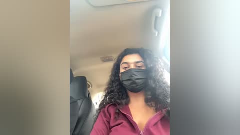 Media: Video of a South Asian woman with long, curly black hair and a medium complexion wearing a black face mask, maroon hoodie, and black seatbelt, seated in a car.