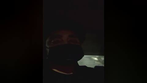 Media: A dimly lit video of a man in a dark mask, standing in a shadowy room with a faint light source on the left. The background is indistinct and dark, enhancing the mysterious and threatening atmosphere.