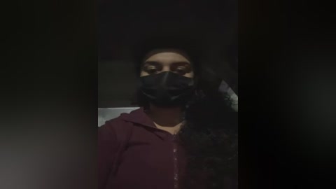 Media: Video of a person with long black hair and dark skin, wearing a black face mask and a dark hoodie, standing in a dimly lit, shadowy room.