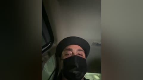 Media: A video of a man with a light skin tone, wearing a black mask, turban, and black clothing, standing in a dark, narrow space with a window to the left.